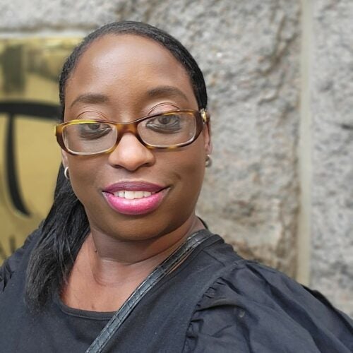 A woman with glasses smiles at the camera. She has long dark hair pulled back into a ponytail, brown glasses and pink lipstick. She is wearing a black top and posed outdoors in front of a granite wall.