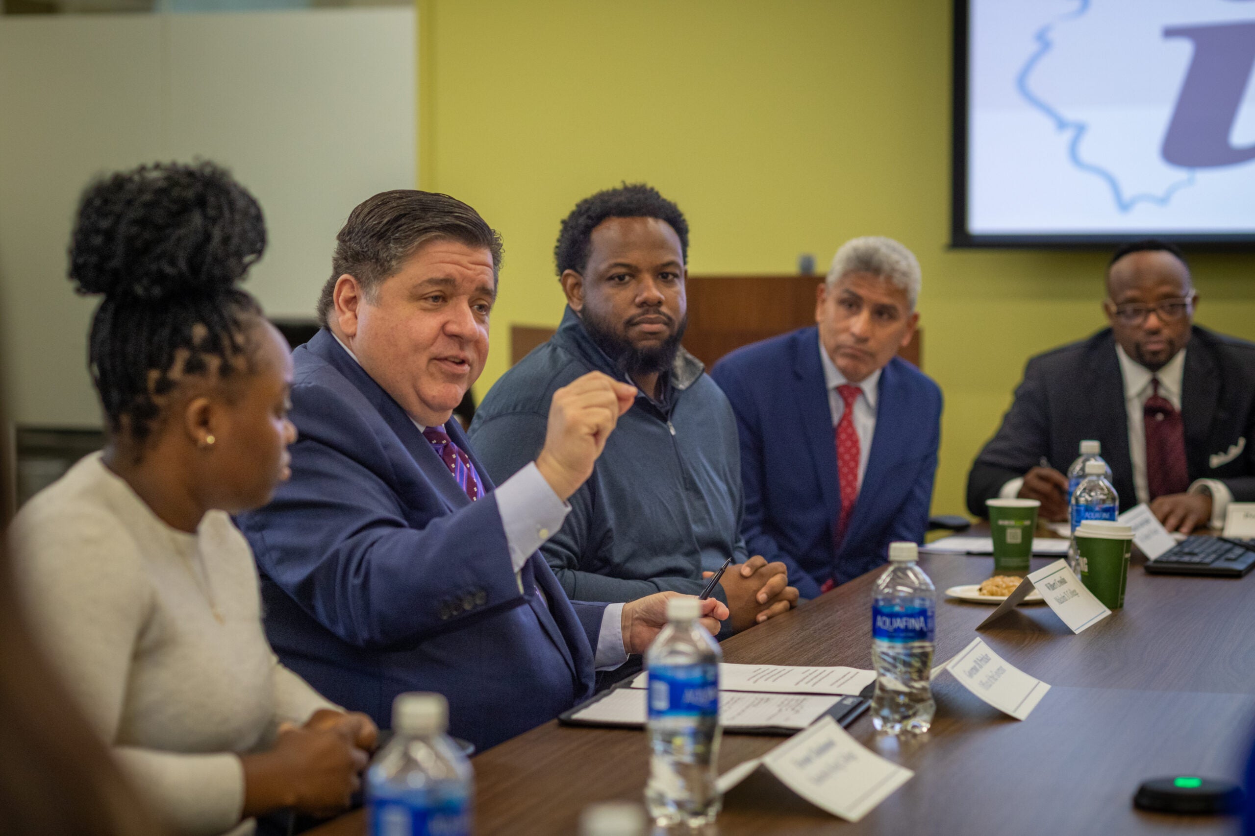 A group of five people sit at a conference table and listen as a man in the middle speaks animatedly.