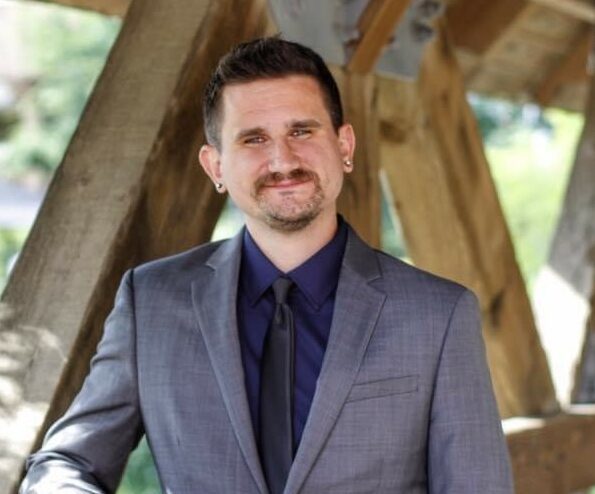 A man in a gray suit coat with a black tie and navy blue shirt smiles at the camera. He has brown hair, a brown mustache and a brown goatee.